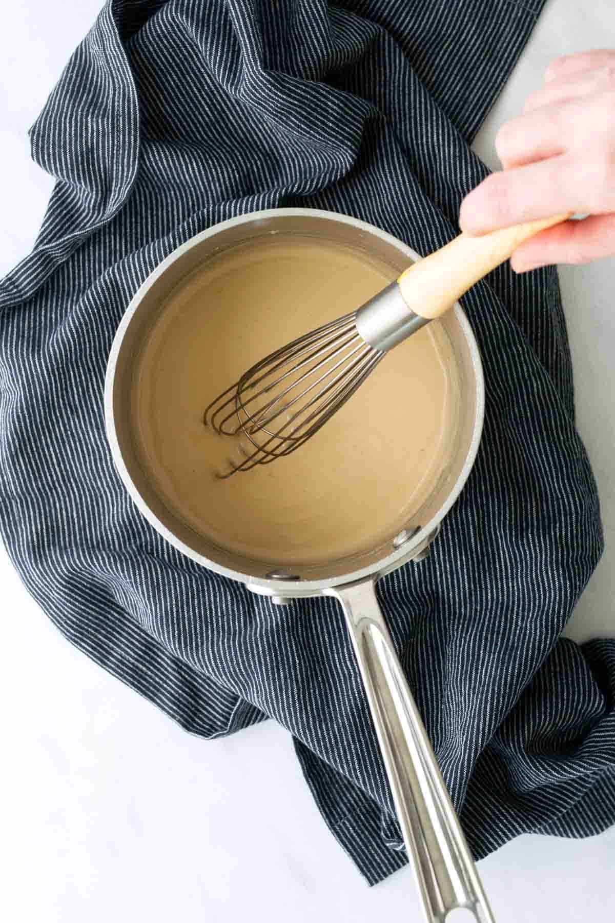 A hand whisking a light brown sauce in a saucepan placed on a dark striped cloth.