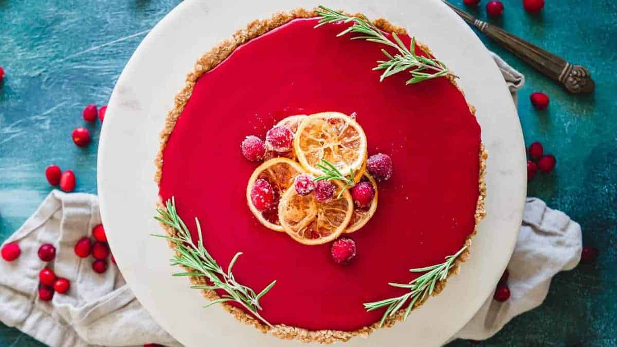 A round tart with a glossy red filling, garnished with candied orange slices, cranberries, and sprigs of rosemary, on a white plate.