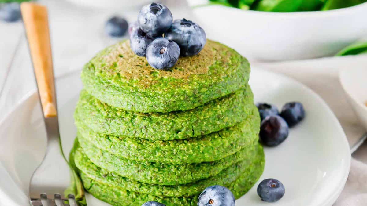 A stack of green pancakes topped with blueberries is served on a white plate with extra blueberries and a fork on the side.
