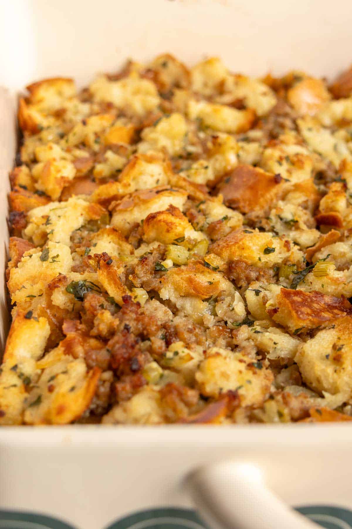 A close-up of baked bread stuffing with herbs and browned sausage pieces in a white casserole dish.