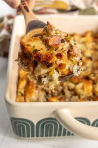 A wooden spoon holds a serving of baked stuffing with bread cubes, vegetables, and herbs above a casserole dish filled with more stuffing.