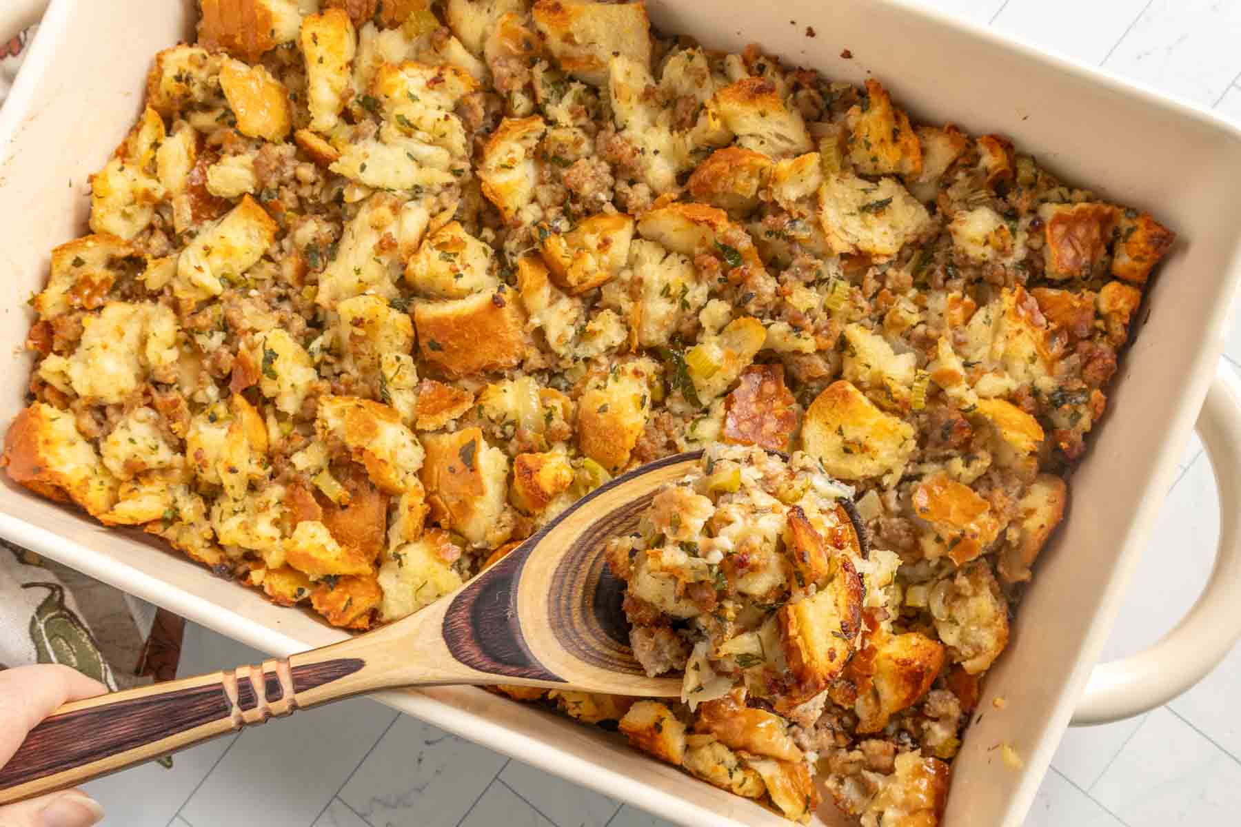 A baking dish filled with bread stuffing mixed with herbs and sausage, with a wooden spoon lifting a portion from the dish.