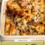 A close-up of a serving spoon lifting sausage stuffing with fresh herbs from a baking dish. The stuffing contains bread, herbs, and sausage.