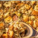 A wooden spoon lifts a serving of sausage stuffing made with bread cubes and herbs from a baking dish.