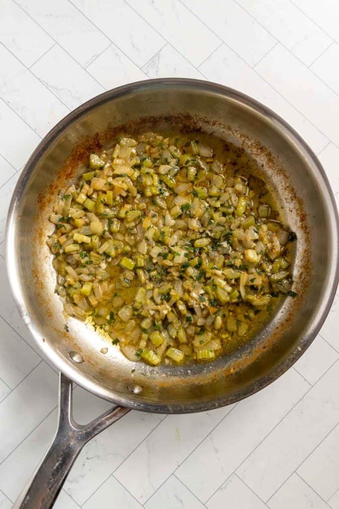 Chopped onions and celery sautéing in a stainless steel pan on a white tiled surface.