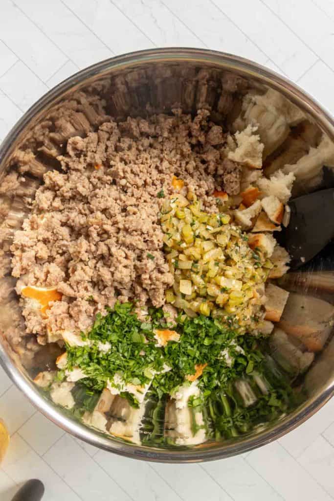 A mixing bowl containing ground meat, chopped cooked vegetables, bread pieces, and fresh herbs, ready to be combined for a recipe.