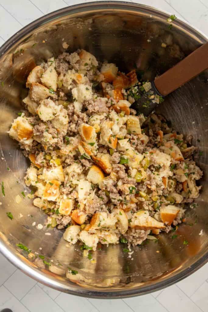 A large metal mixing bowl filled with a stuffing mixture of bread cubes, ground sausage, chopped herbs, and vegetables, being stirred with a spatula.