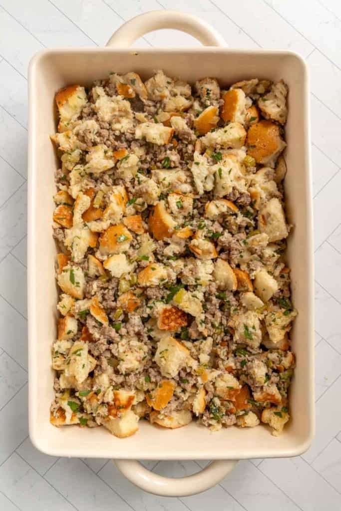 Rectangular baking dish filled with bread stuffing mixed with ground meat, herbs, and small vegetable pieces on a light surface.