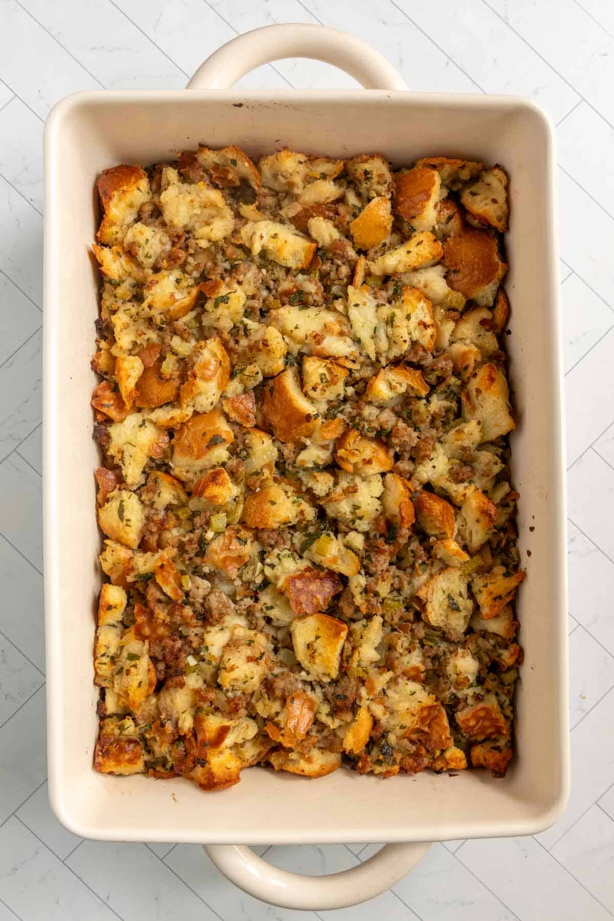 A rectangular baking dish filled with baked bread stuffing mixed with herbs and browned sausage pieces.
