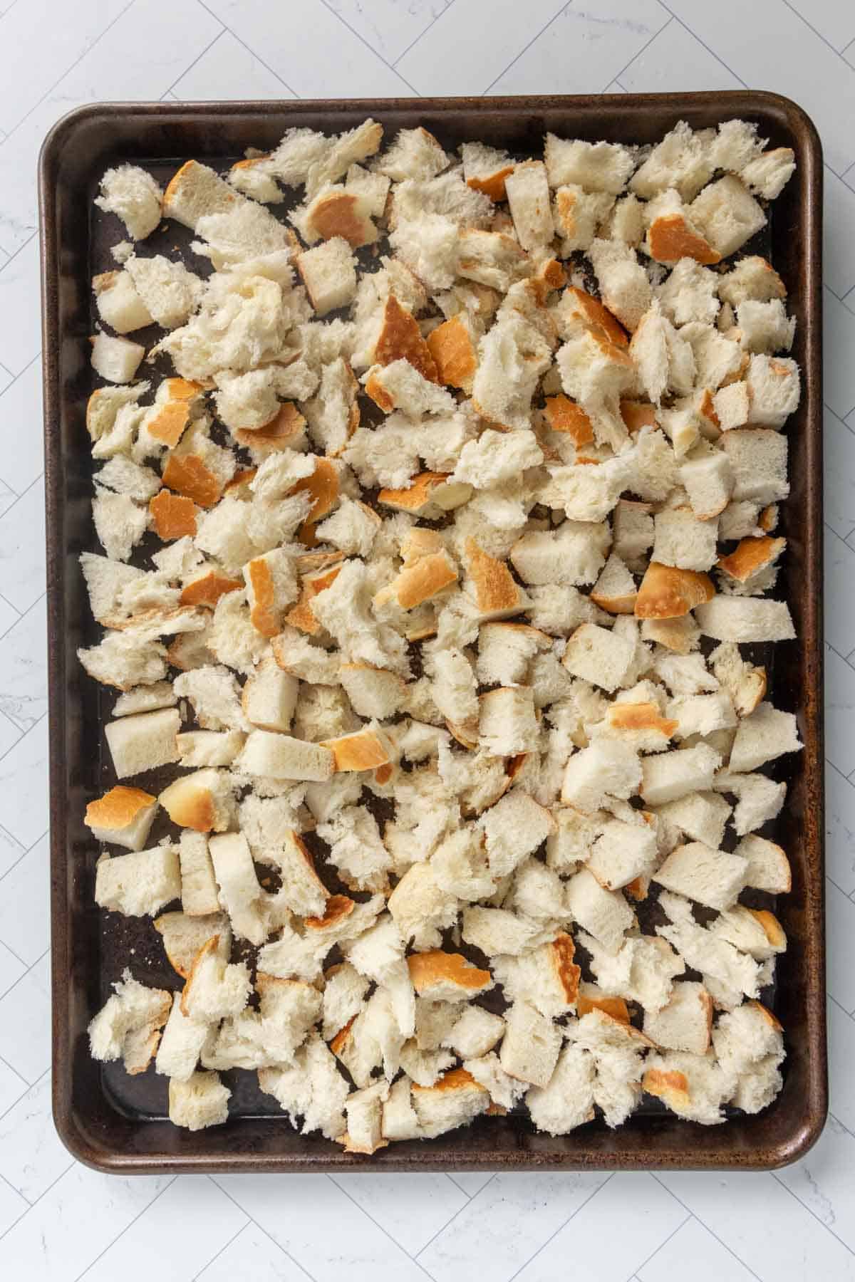 A baking sheet covered with unevenly torn pieces of white bread, spread out in a single layer on a light-colored surface.
