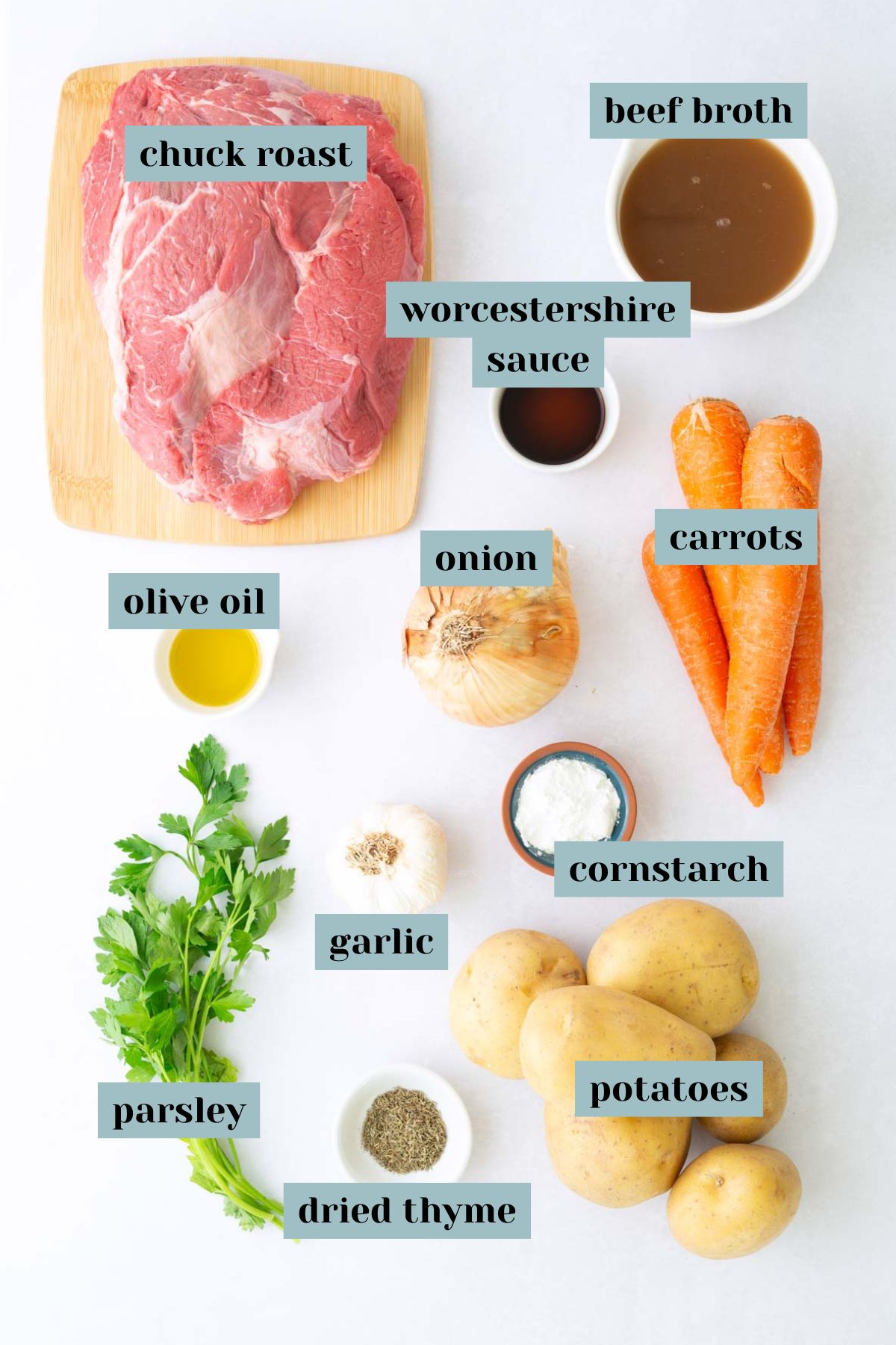 Ingredients for a beef stew recipe arranged on a white surface, including chuck roast, broth, carrots, potatoes, onion, garlic, parsley, dried thyme, olive oil, Worcestershire sauce, and cornstarch.