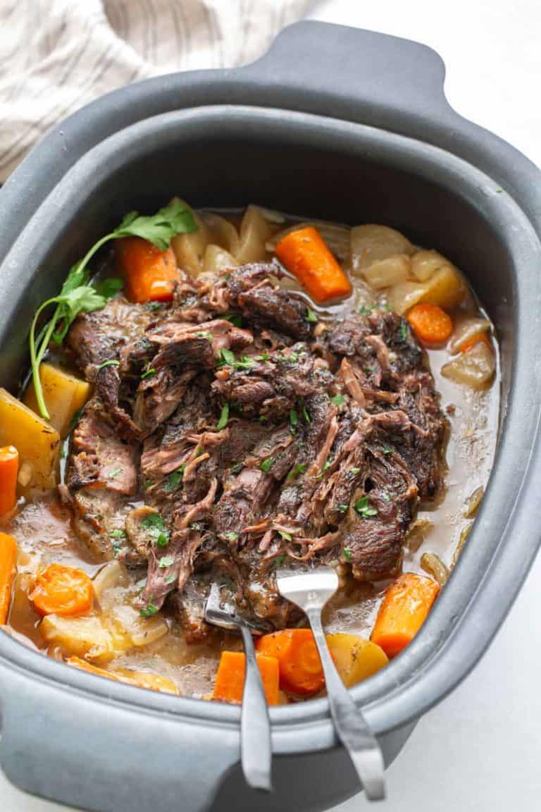 A pot roast with shredded beef, carrots, potatoes, and onions in a slow cooker, garnished with fresh herbs, with two forks resting in the meat.