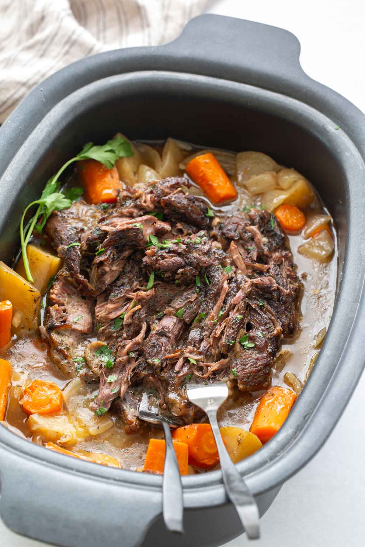 A pot roast with shredded beef, carrots, potatoes, and onions in a slow cooker, garnished with fresh herbs, with two forks resting in the meat.