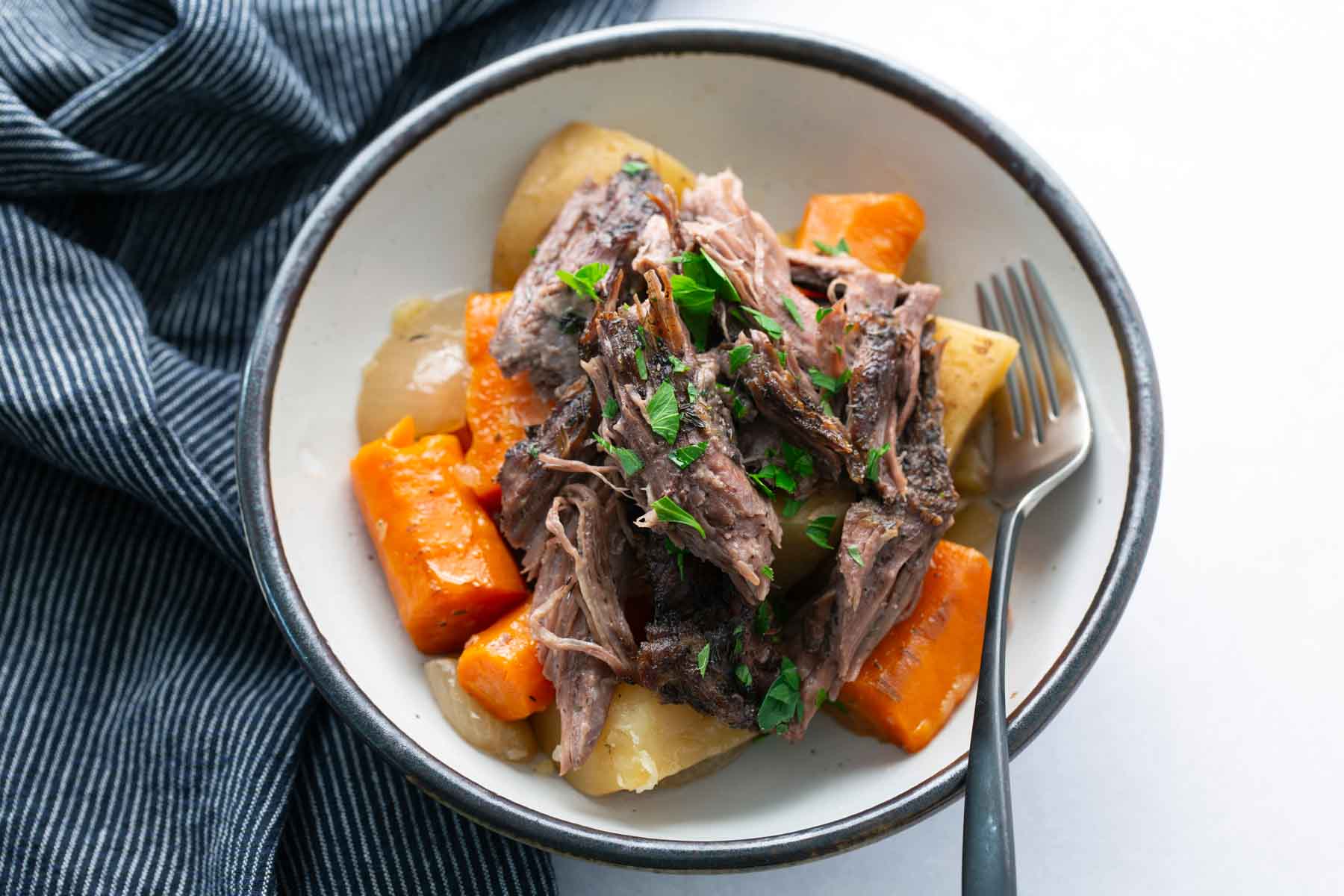 A bowl of pot roast with shredded beef, carrots, and potatoes, garnished with chopped parsley, next to a fork and a blue striped cloth.
