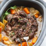 A slow cooker filled with pot roast, surrounded by chunks of potatoes, carrots, and onions in broth, with two serving forks in the meat.