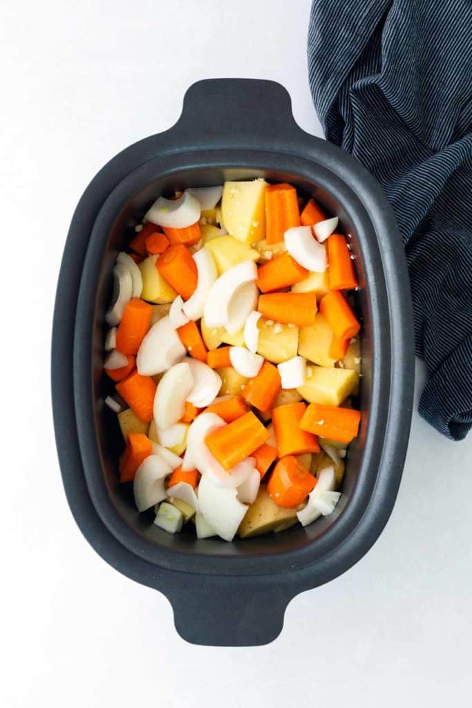 Chopped potatoes, carrots, and onions in a black slow cooker, ready to be cooked, with a dark cloth on the side.