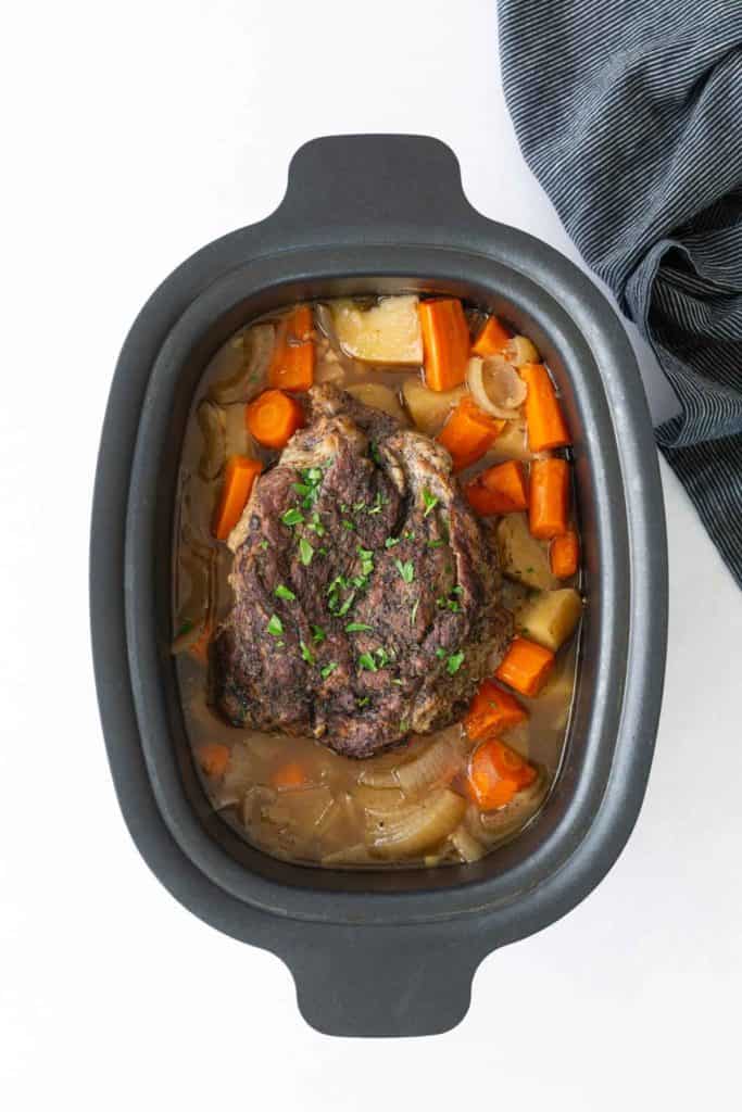 A cooked pot roast with carrots, potatoes, and onions in a black roasting pan, garnished with chopped parsley.