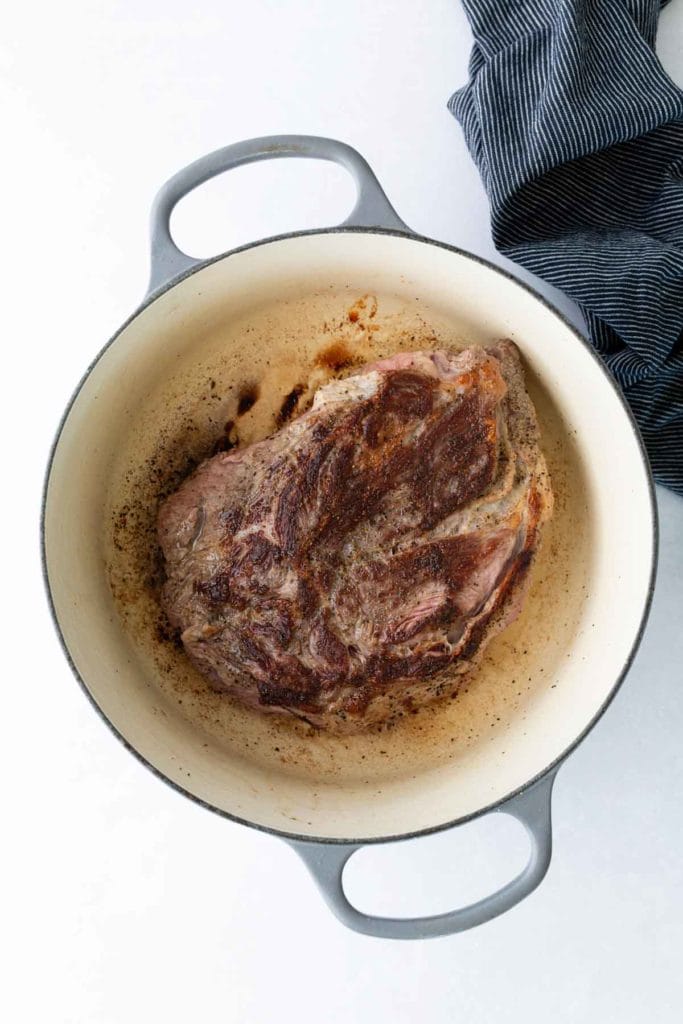 A browned piece of meat sits in a large, round, white Dutch oven with a dark rim. A striped cloth is partially visible in the upper right corner.