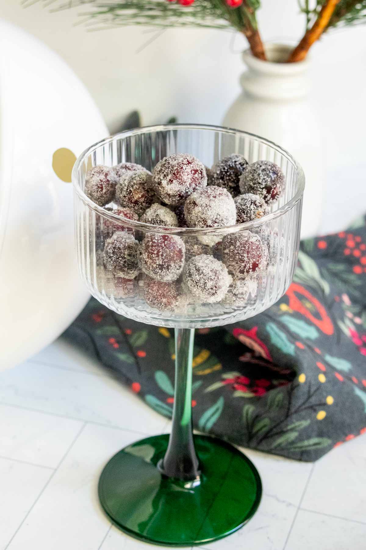 A clear glass goblet filled with sugared cranberries sits on a white surface with a black patterned napkin and a white vase in the background.