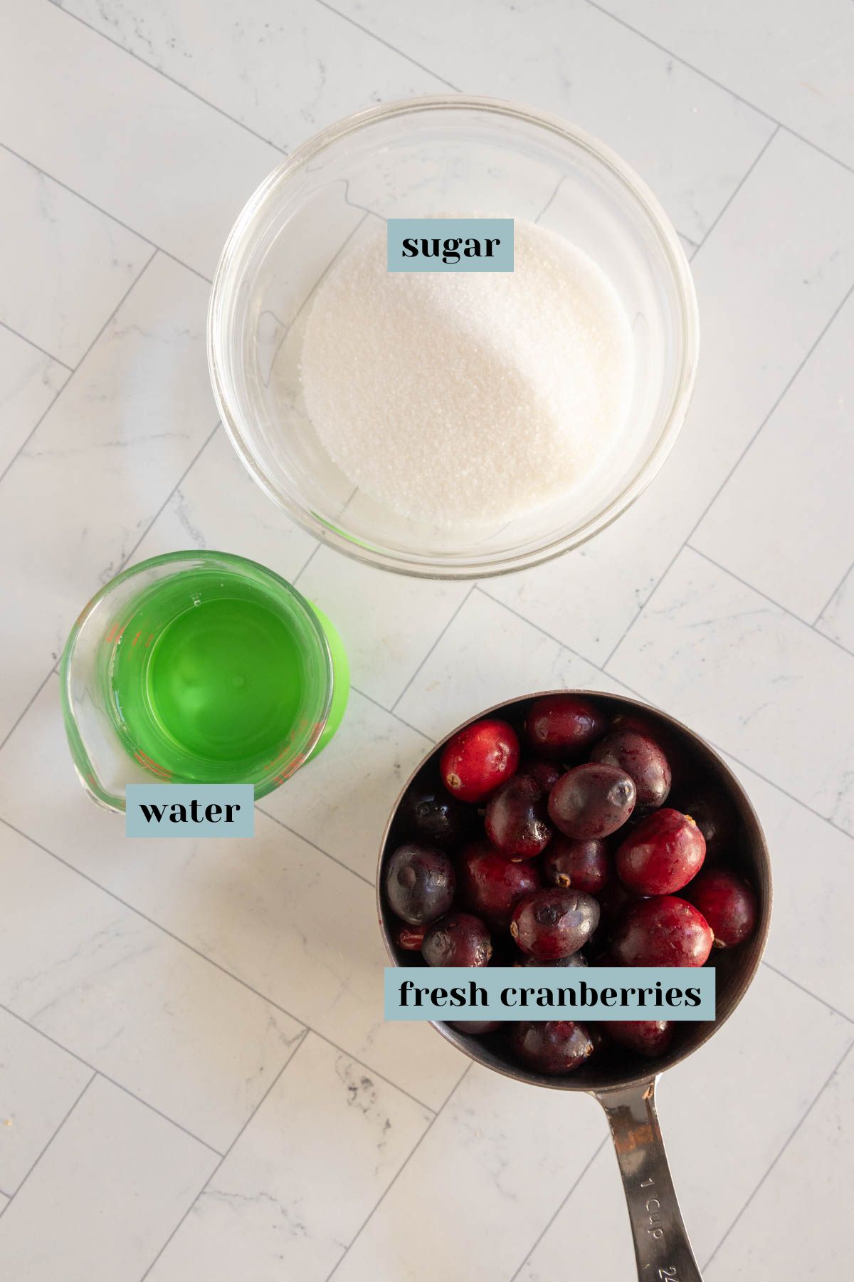 A bowl of sugar, a cup of water, and a measuring cup of fresh cranberries are arranged on a white countertop, each labeled accordingly.
