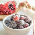 A white bowl filled with sugared cranberries, with a red mesh bag of fresh cranberries and a decorated cloth in the background.