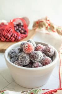 A white bowl filled with sugared cranberries, with a red mesh bag of fresh cranberries and a decorated cloth in the background.