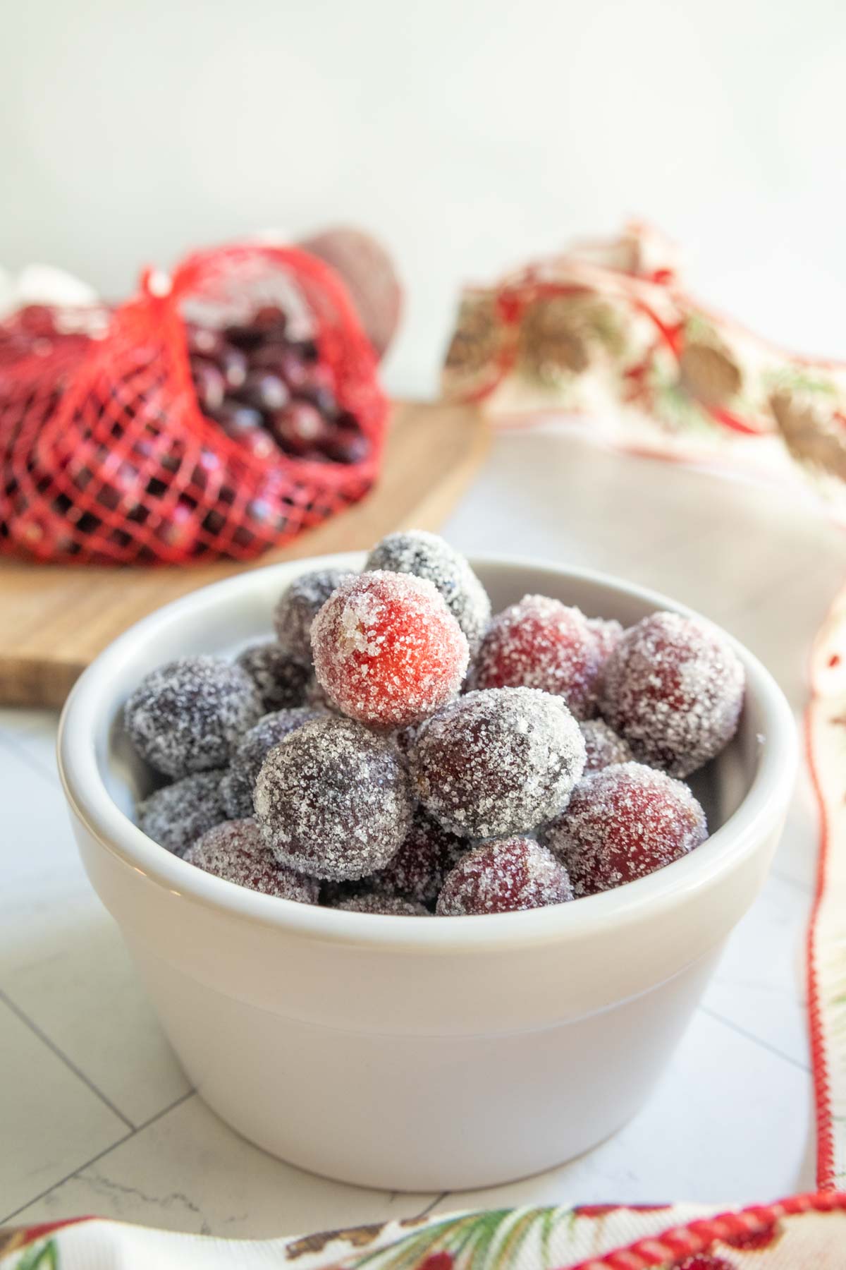A white bowl filled with sugared cranberries, with a red mesh bag of fresh cranberries and a decorated cloth in the background.