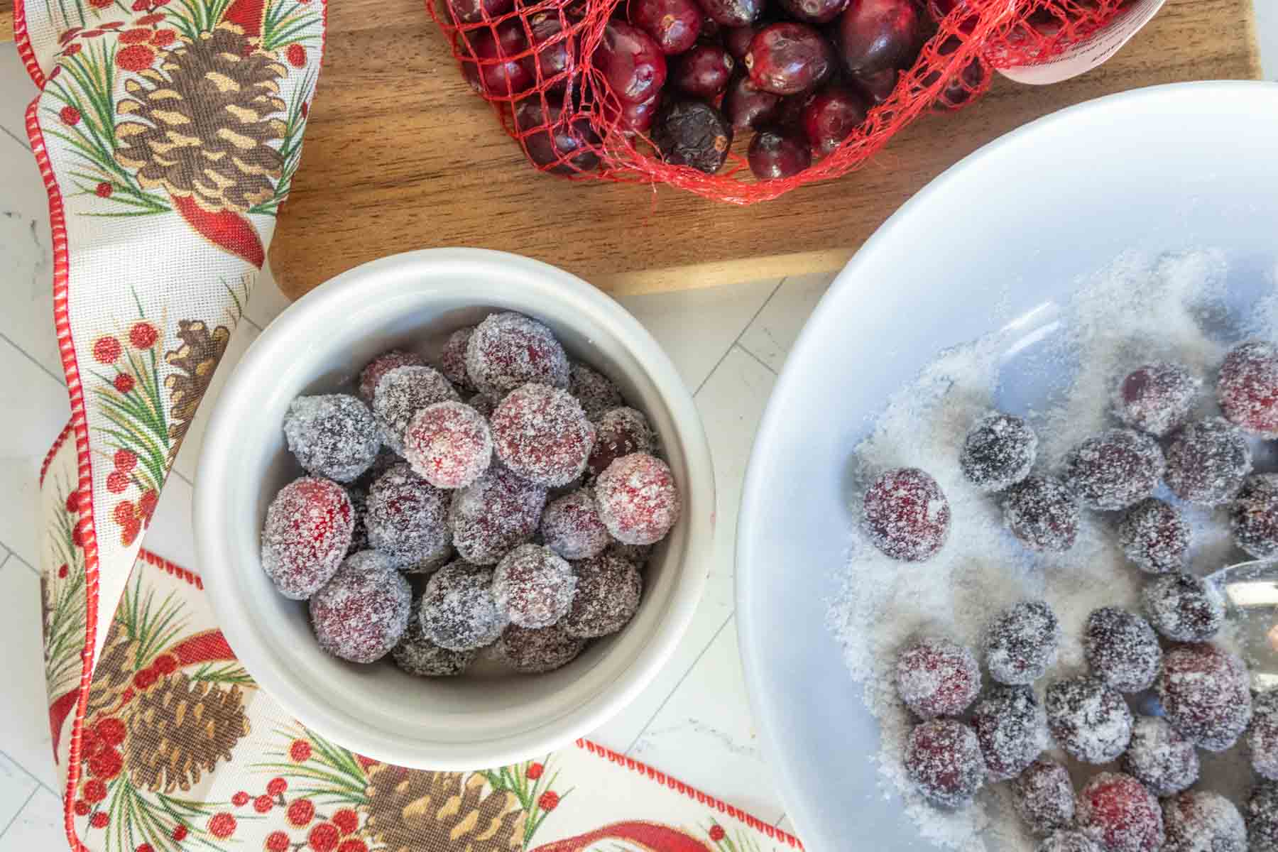 A bowl of sugared cranberries sits next to a bowl of cranberries being coated in sugar, a bag of fresh cranberries, and a festive holiday ribbon with pinecone and holly designs.