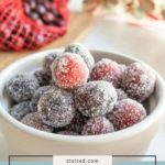 A white bowl filled with sugared cranberries sits on a counter; a bag of fresh cranberries is in the background.