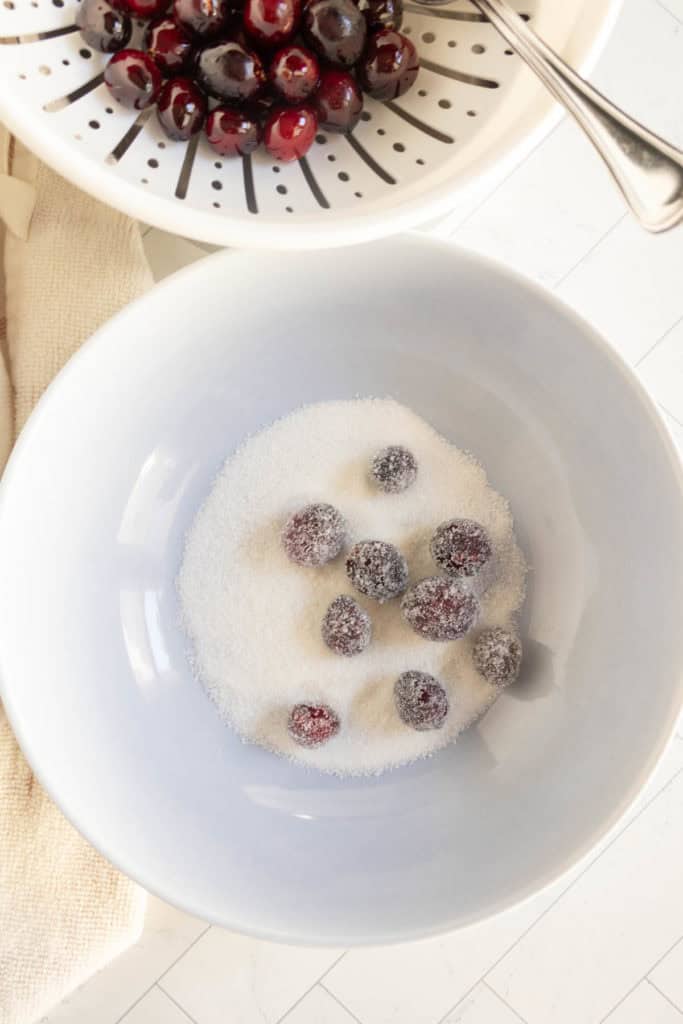 A white bowl with cranberries being coated in sugar, next to a colander containing rinsed cranberries on a white surface.