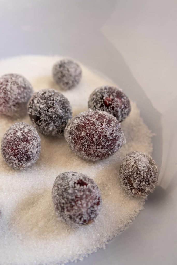 Several cranberries coated in granulated sugar rest on a bed of sugar against a neutral background.