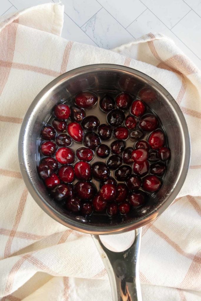 A saucepan filled with whole cherries in liquid sits on a beige and white checkered cloth on a tiled surface.