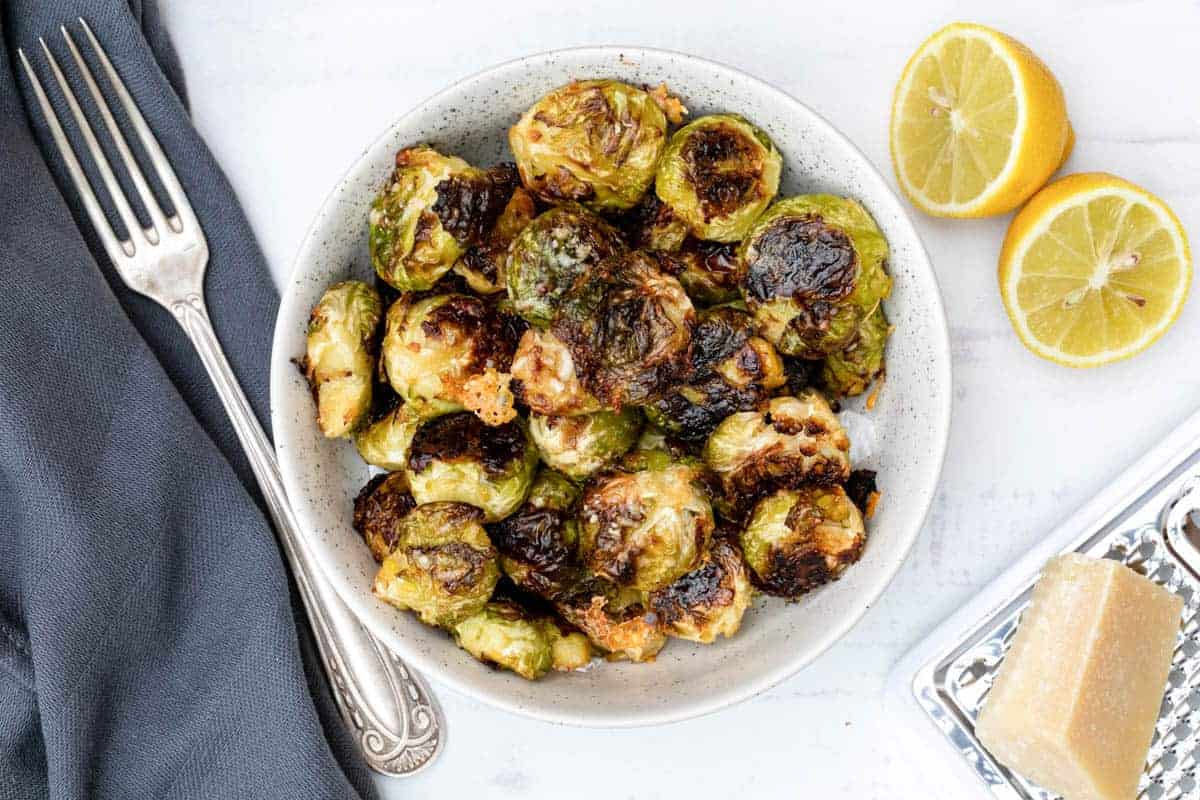 A bowl of roasted Brussels sprouts with a fork, two lemon halves, a block of cheese, and a gray napkin on a white surface.