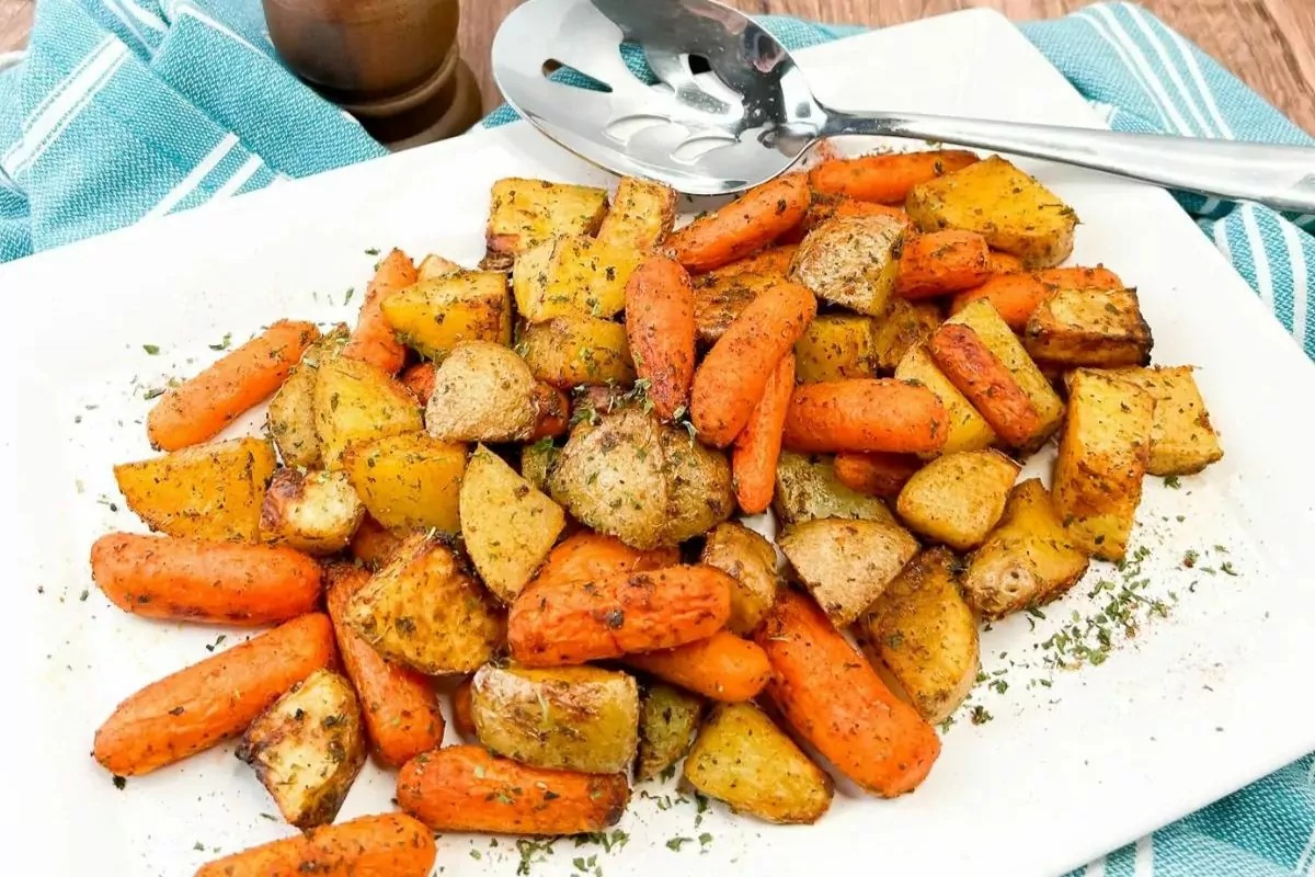 A white plate filled with roasted potatoes and baby carrots, garnished with herbs, next to a serving utensil on a blue striped cloth.