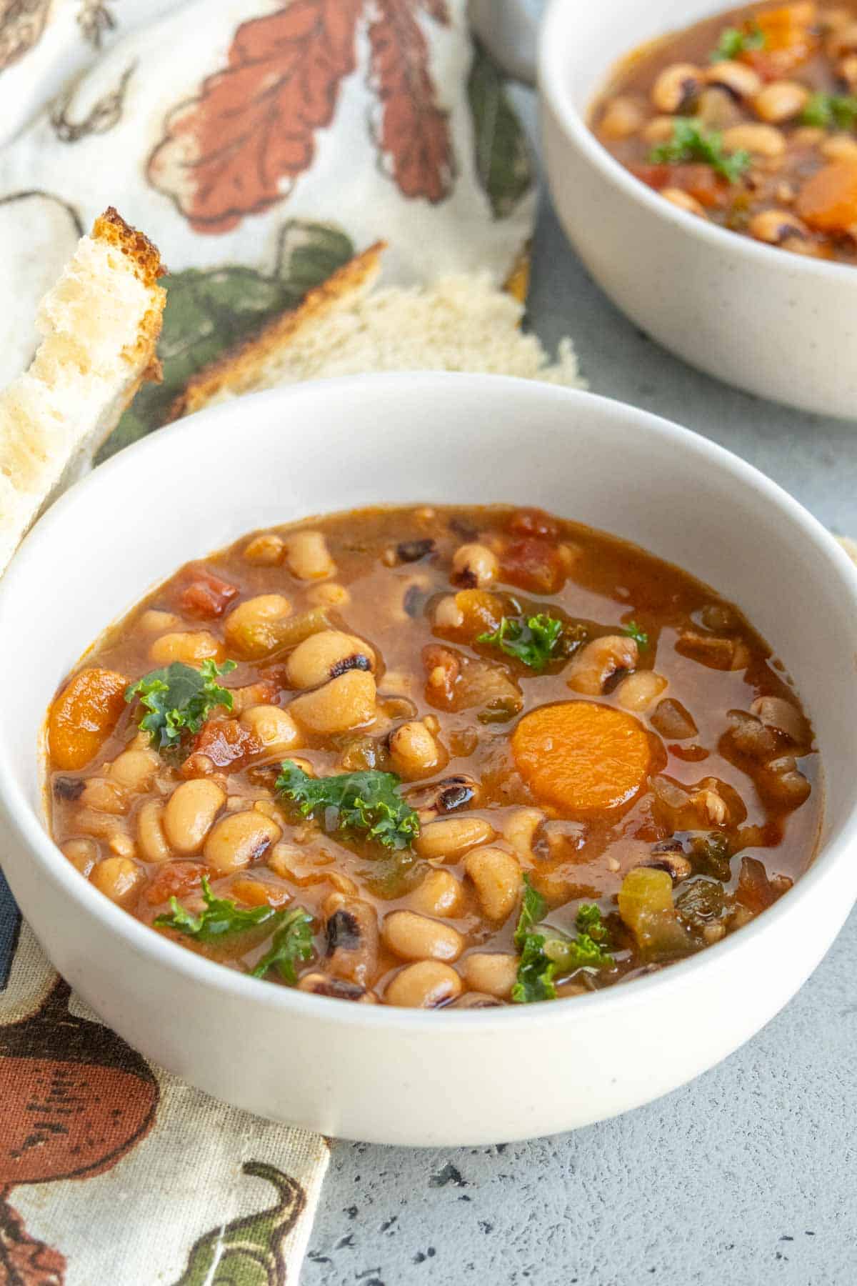 A white bowl filled with vegetable and bean soup, featuring black-eyed peas, carrots, greens, and tomatoes, on a table with bread and a patterned napkin.