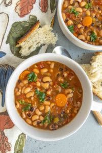 Two bowls of black-eyed pea soup with carrots and herbs, served with slices of bread on the side, placed on a patterned cloth.