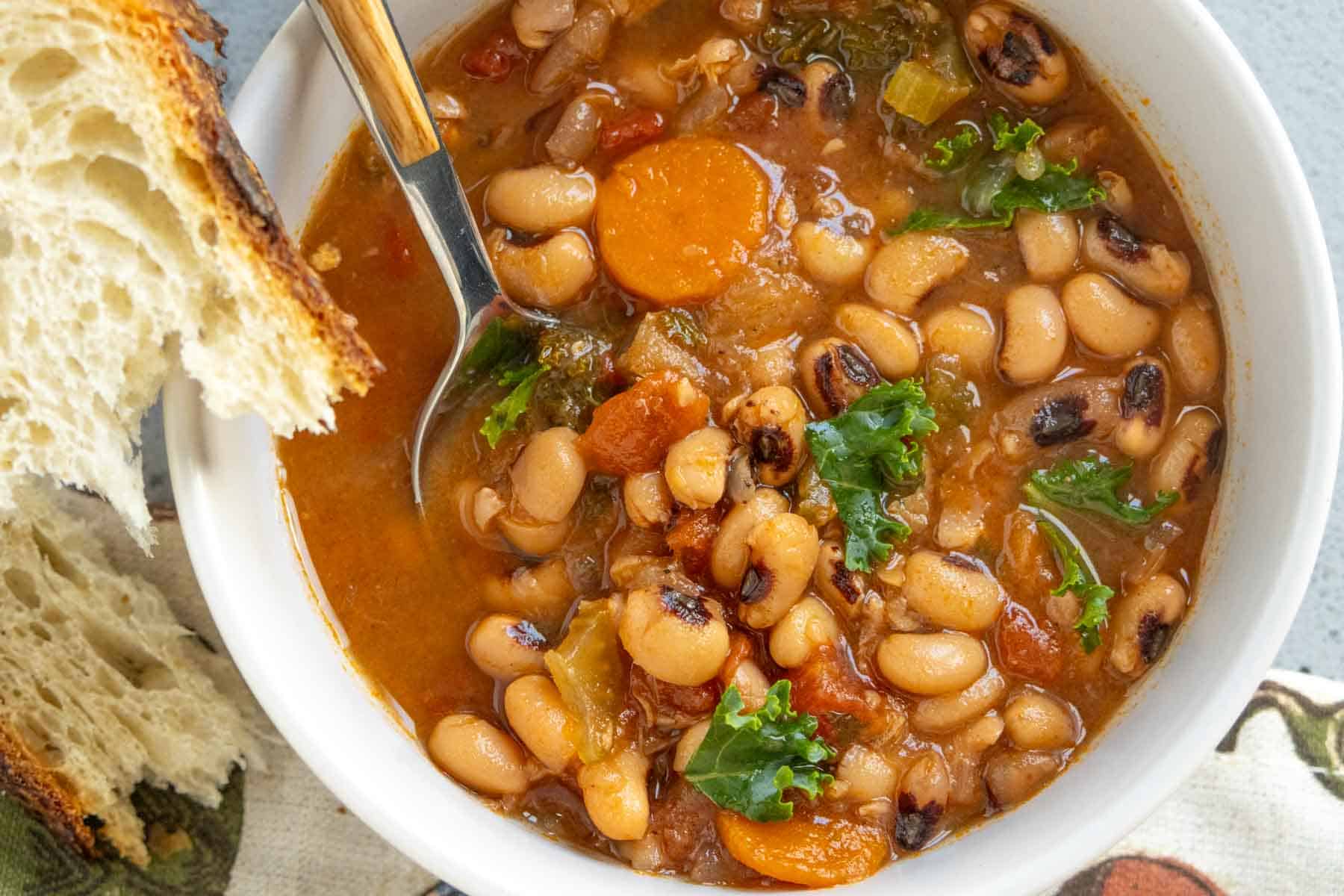 A bowl of black-eyed pea soup with carrots, greens, and tomatoes, served with a slice of rustic bread and a spoon.