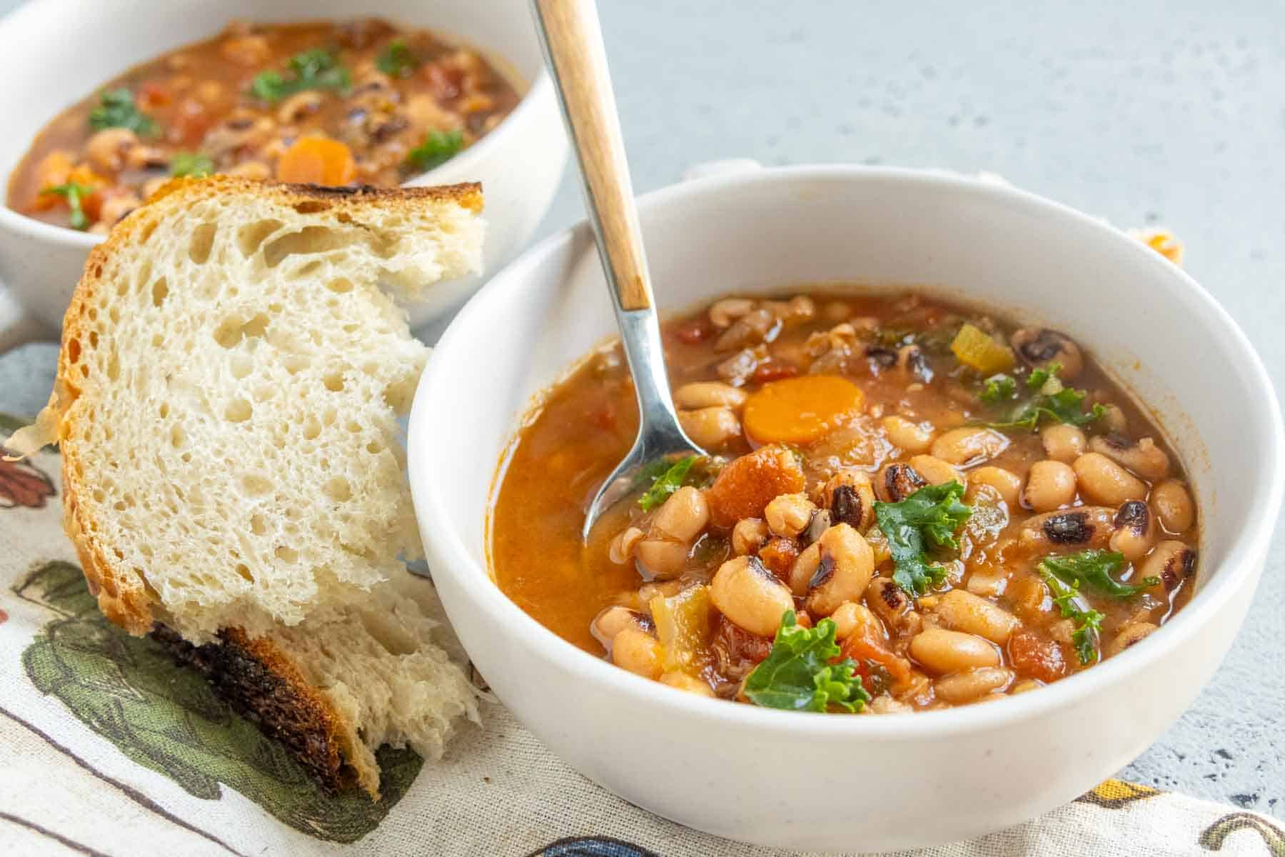 A bowl of bean and vegetable soup with a spoon, served with a slice of rustic bread. Another bowl of soup is visible in the background.