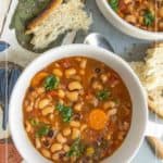 Two bowls of black-eyed pea soup with carrots and herbs, served with slices of rustic bread on a table with a patterned cloth.