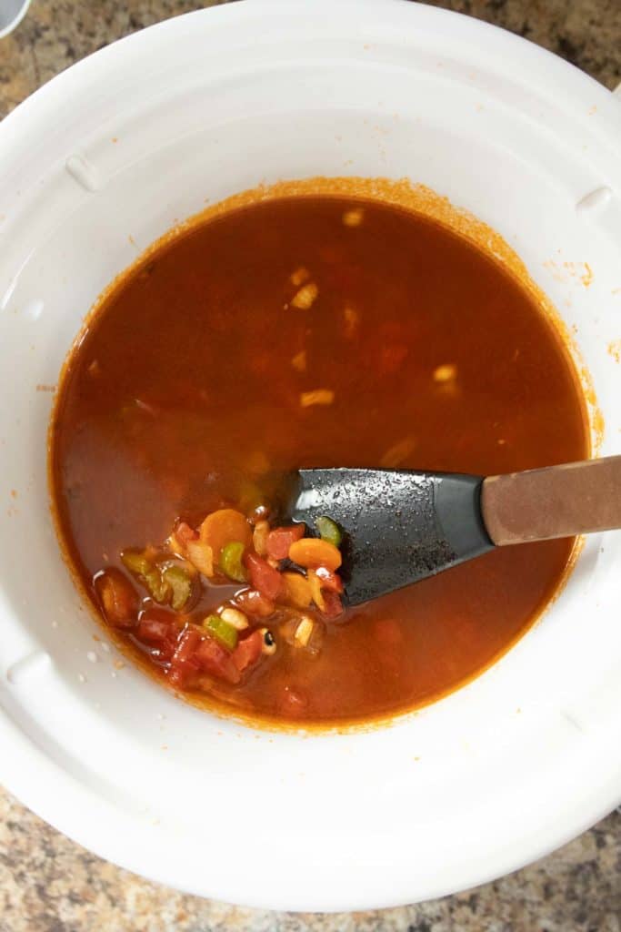 A white slow cooker filled with a tomato-based vegetable soup, featuring chopped carrots, celery, and tomatoes, being stirred with a black spatula.