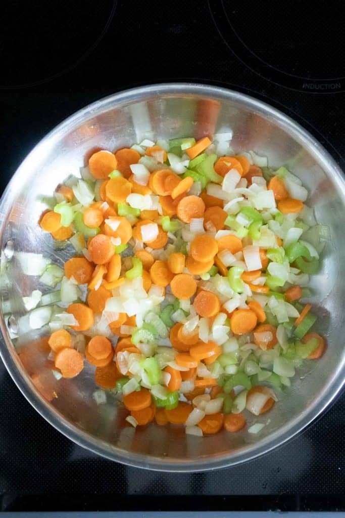 Chopped carrots, celery, and onions cooking in a stainless steel pan on a stovetop.