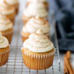 Pumpkin cupcakes with piped cream cheese frosting are arranged on a cooling rack, with cinnamon sticks and a dark cloth in the background.