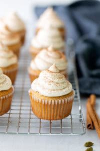 Pumpkin cupcakes with piped cream cheese frosting are arranged on a cooling rack, with cinnamon sticks and a dark cloth in the background.