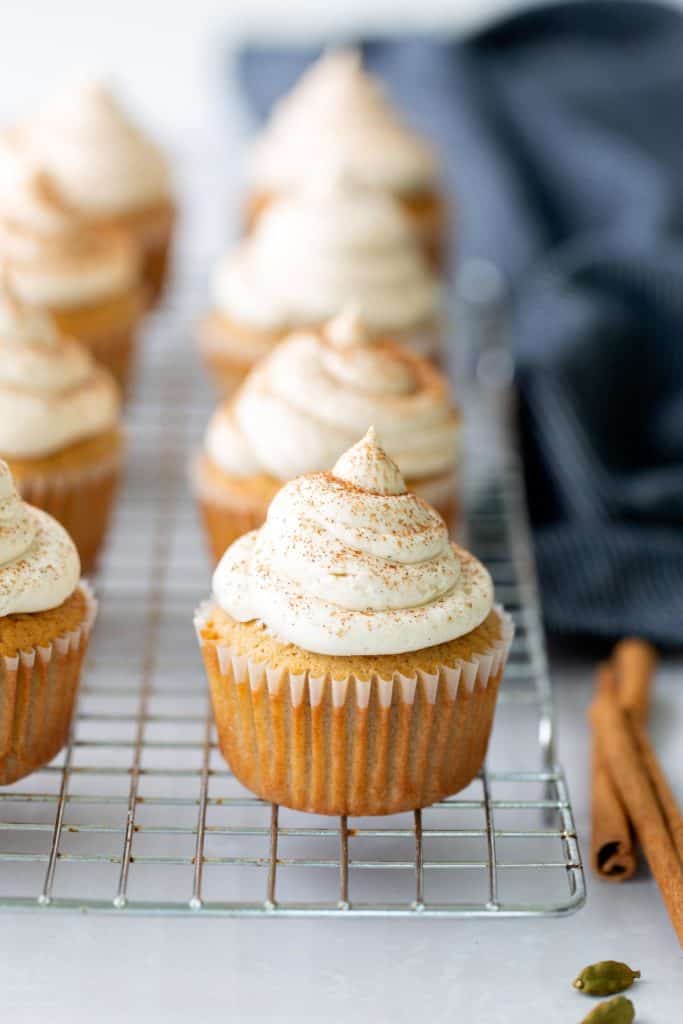 Chai Cupcakes Pumpkin cupcakes with piped cream cheese frosting are arranged on a cooling rack, with cinnamon sticks and a dark cloth in the background.