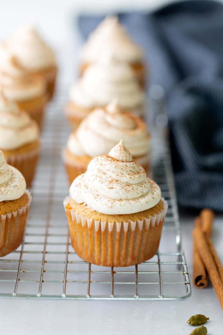 Pumpkin cupcakes with piped cream cheese frosting are arranged on a cooling rack, with cinnamon sticks and a dark cloth in the background.
