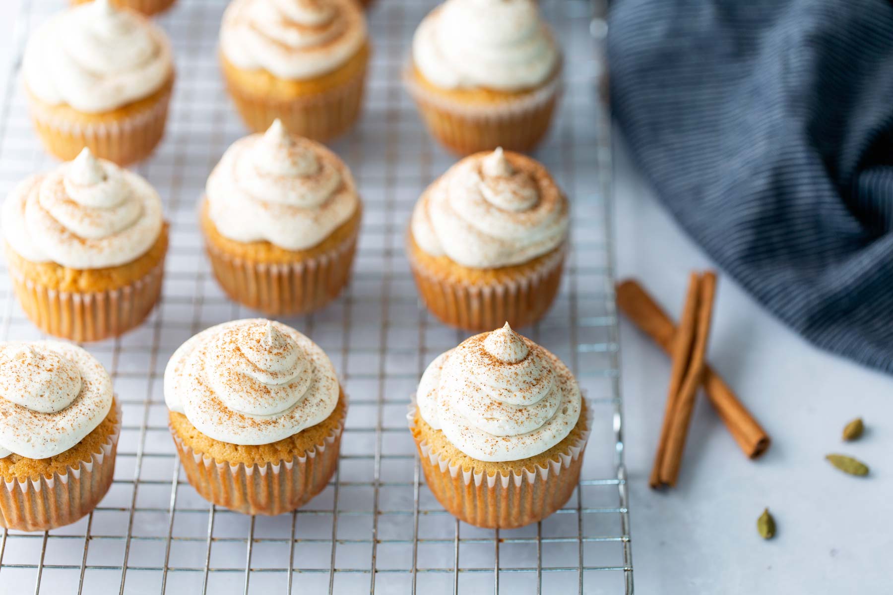 Nine cupcakes with swirled frosting and cinnamon dusting are arranged on a cooling rack, with cinnamon sticks and a striped cloth beside them.