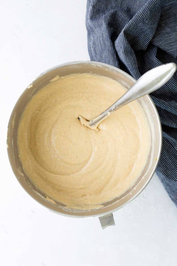A mixing bowl filled with light brown cake batter and a metal spoon, next to a blue and white striped cloth.