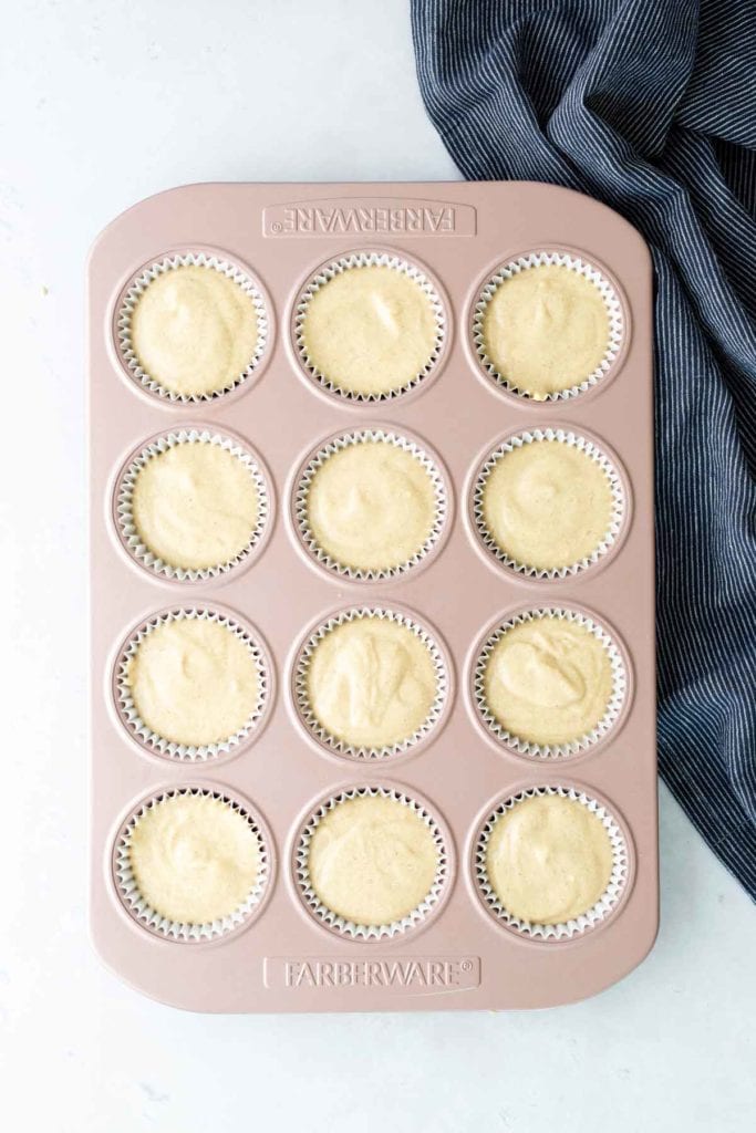 A muffin tin filled with cupcake liners containing unbaked batter sits on a light surface next to a dark striped cloth.