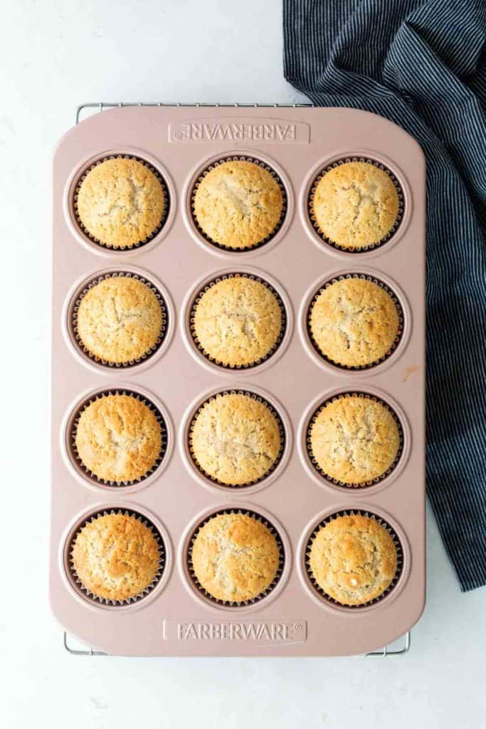 A muffin tin filled with twelve baked muffins, each in a brown paper liner, sits on a light surface next to a folded dark striped cloth.