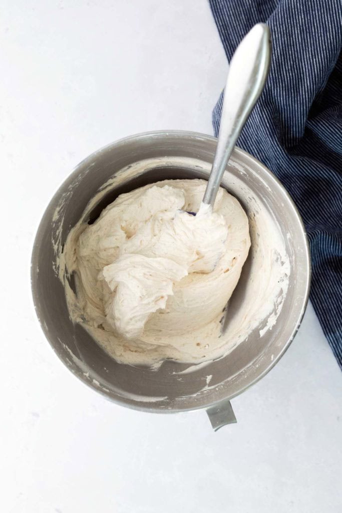 A metal mixing bowl filled with thick, whipped cream or frosting, with a metal spoon sticking out. A blue-striped cloth is partially visible to the side.